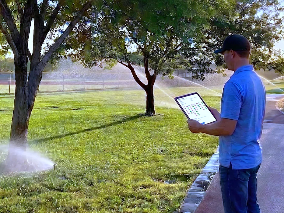 Un técnico registra los hallazgos en una tableta mientras una zona funciona detrás de él, con los aspersores lanzando agua sobre el césped.